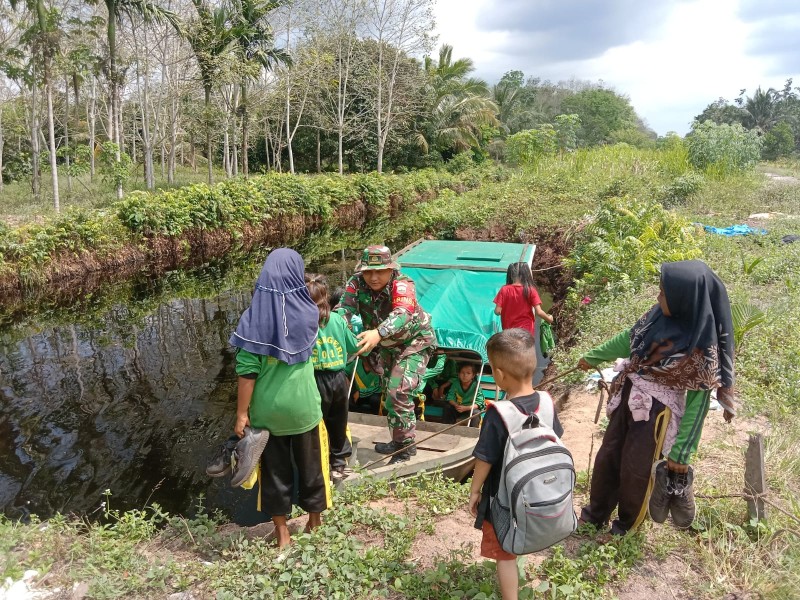 Penuh Dedikasi, Babinsa Koramil 06/Kateman Bantu Anak Sekolah Naik Perahu di Kanal Desa Kelapa Patih Jaya